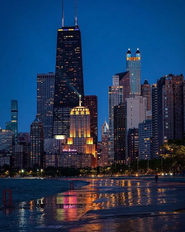 the city skyline is lit up at night, with skyscrapers reflecting in the water