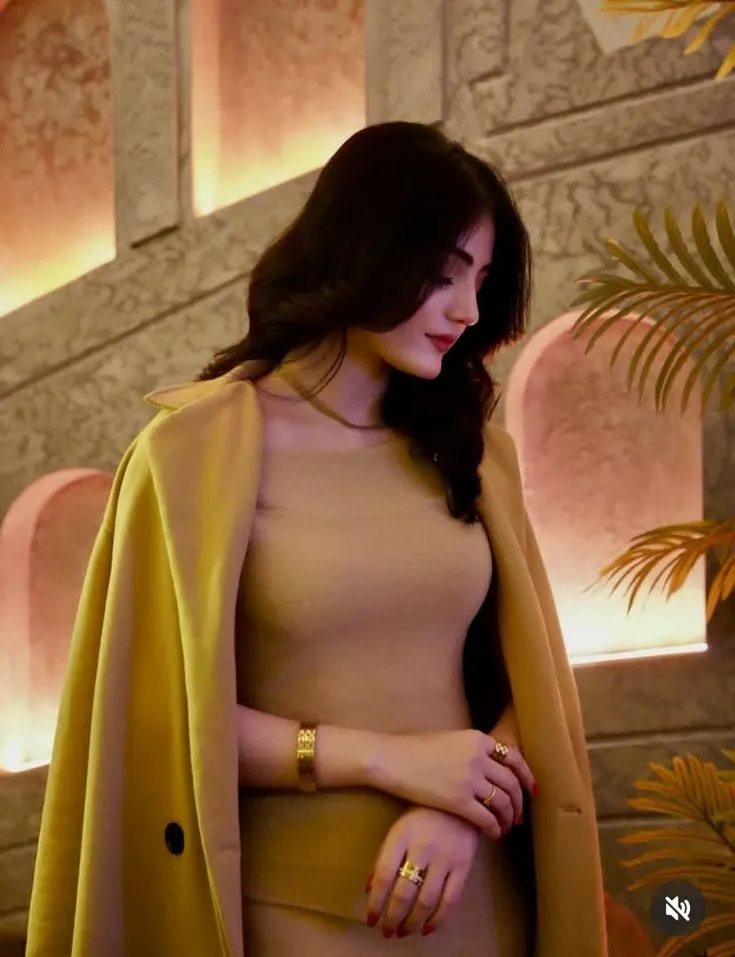 a woman standing in front of a plant wearing a tan dress and yellow jacket with her hands on her hips