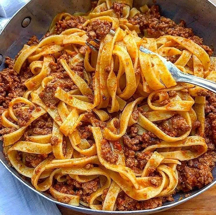 a pan filled with pasta and meat sauce on top of a wooden table next to a fork
