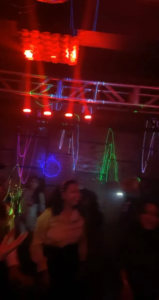 people are dancing in a dark room with lights on the ceiling and some have their arms around each other