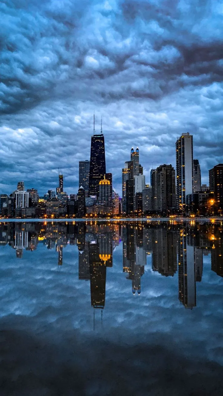 a city skyline is reflected in the water at night with dark clouds and blue sky