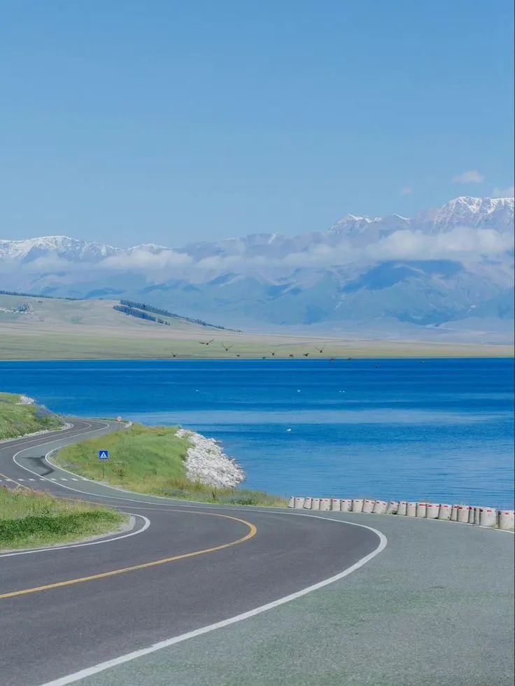 an empty road near the water with mountains in the background