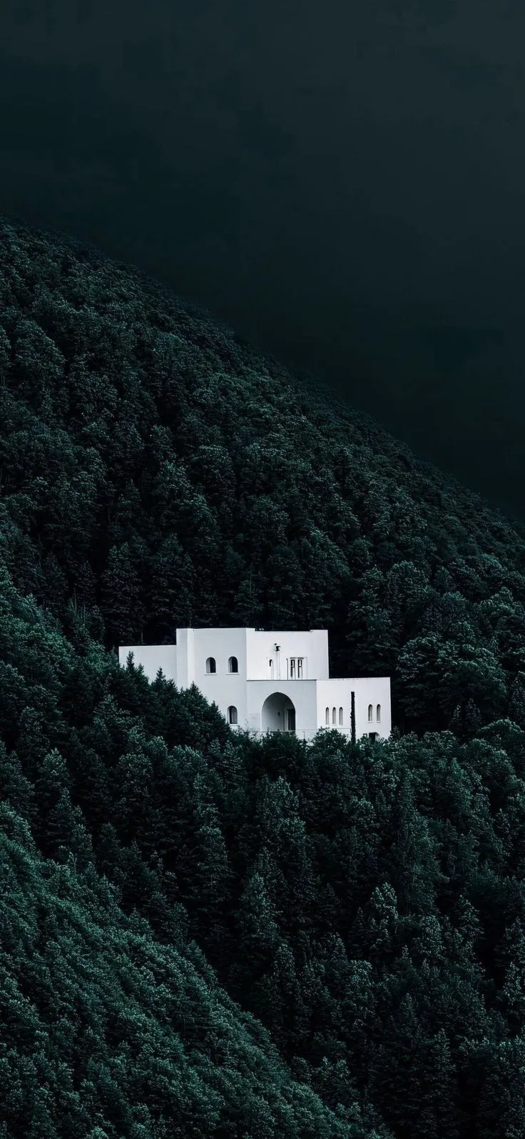 a white house sitting on the side of a lush green forest covered hillside under a dark sky