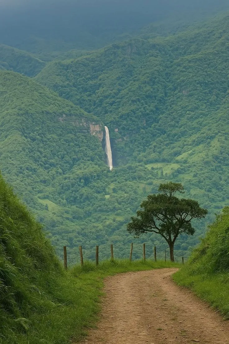 a dirt road leading to a waterfall in the middle of a lush green mountain range