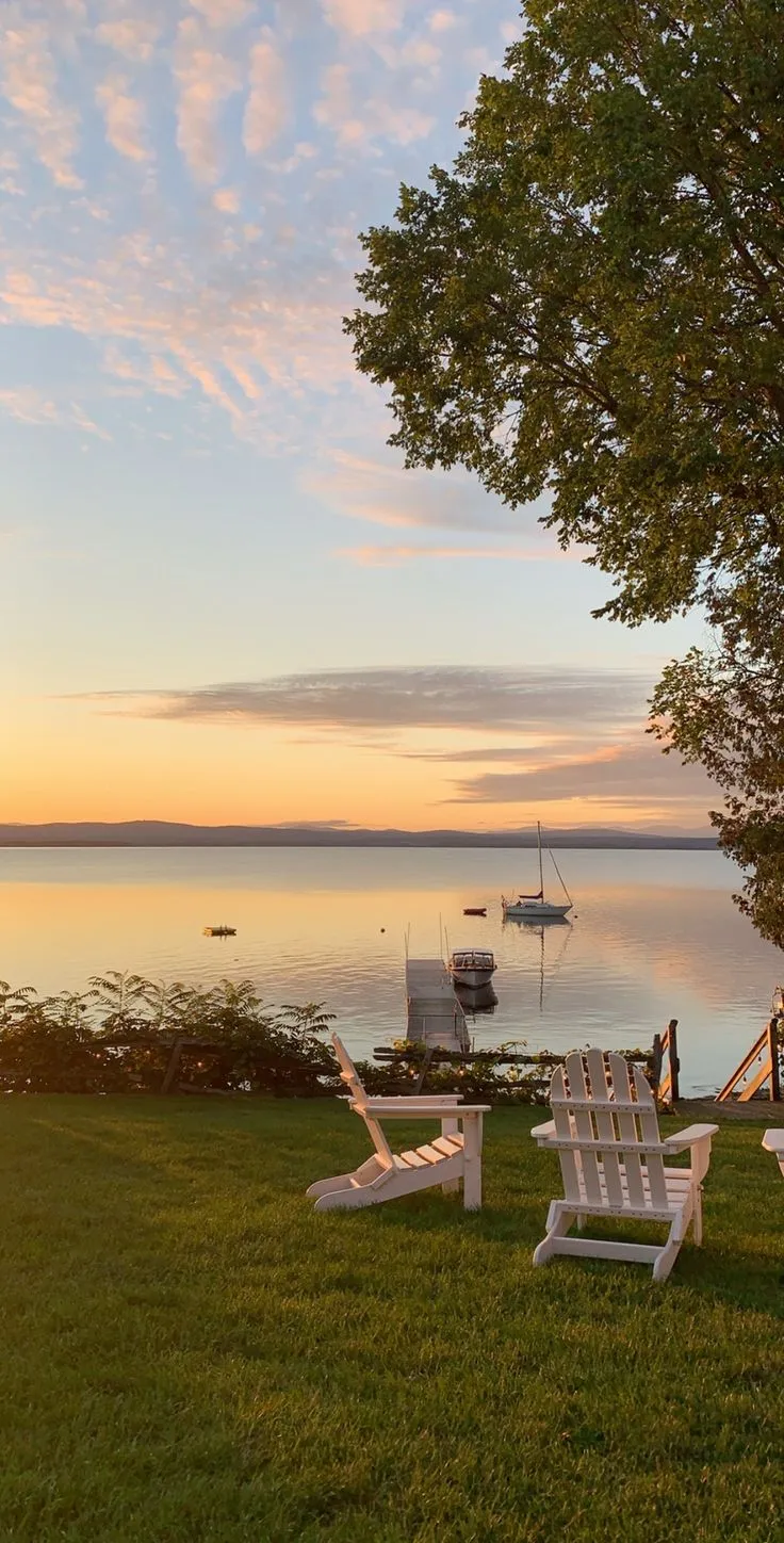 two white lawn chairs sitting on top of a lush green field next to a lake