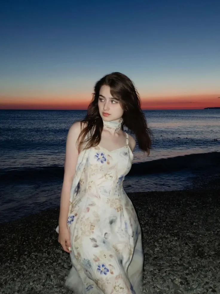a woman in a dress standing on the beach at sunset with her hair blowing in the wind