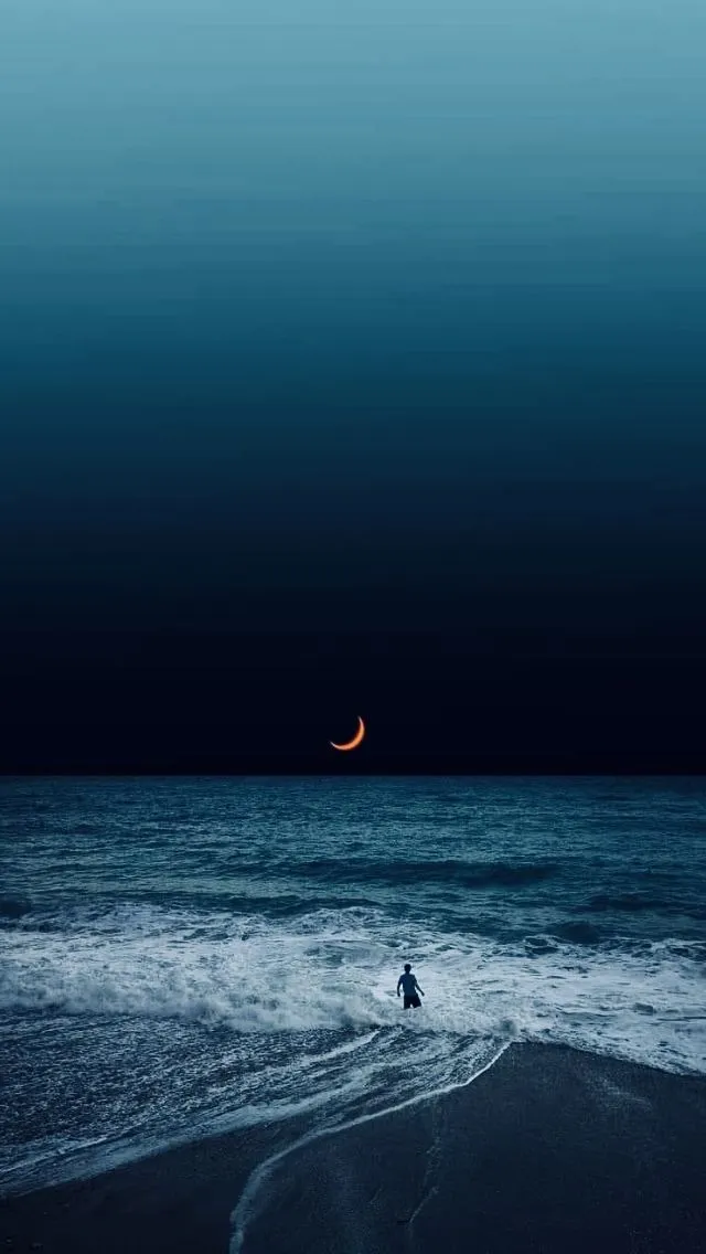 a man standing in the ocean at night with a half moon above him and an orange crescent