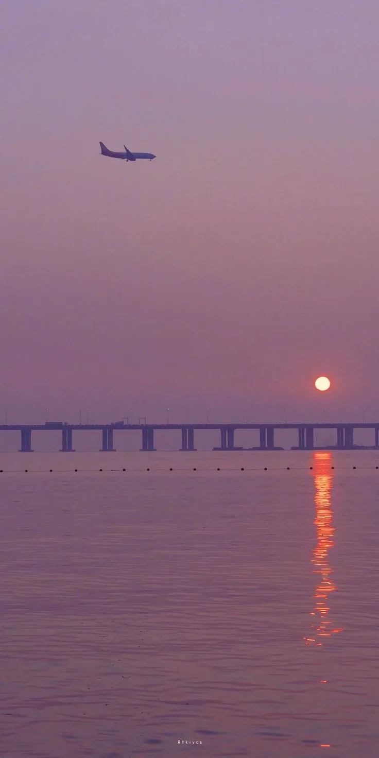 an airplane is flying over the ocean at sunset with a large bridge in the background