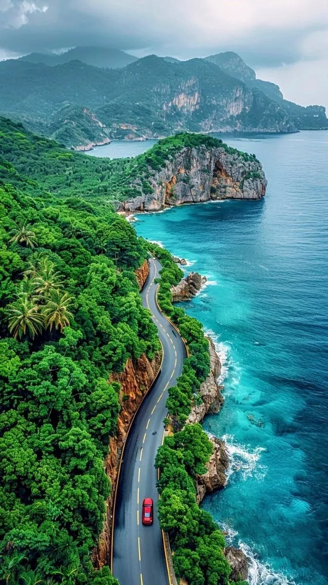 an aerial view of a road winding into the ocean with trees on both sides and mountains in the background