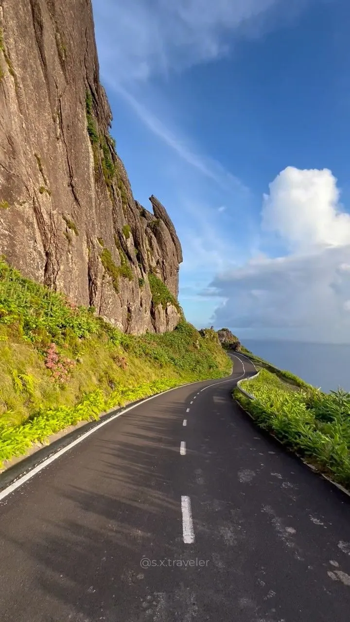 an empty road near the side of a mountain with green vegetation on both sides and blue sky in the background