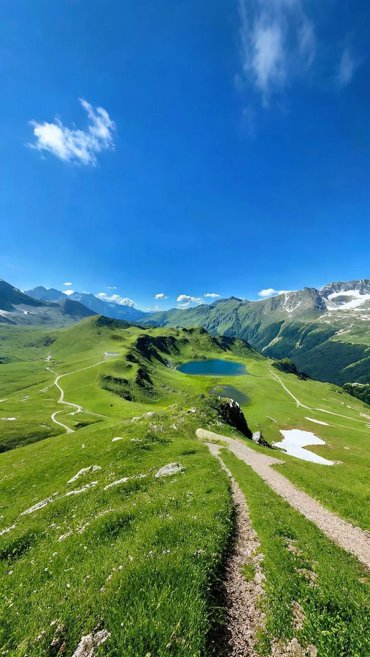 a trail winds through the grass towards a body of water in the distance, surrounded by mountains