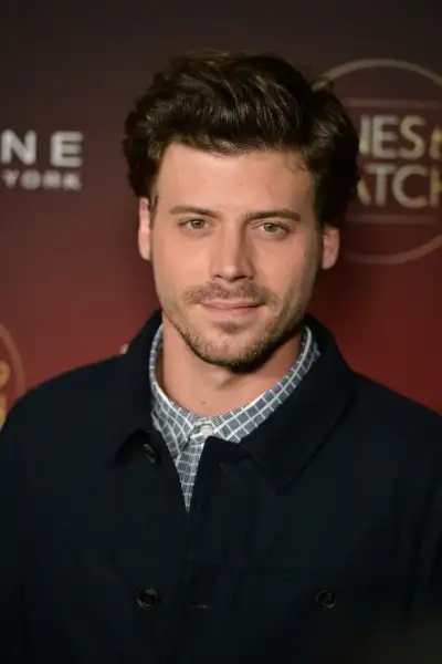 a man in a black coat and plaid shirt posing for a photo on the red carpet