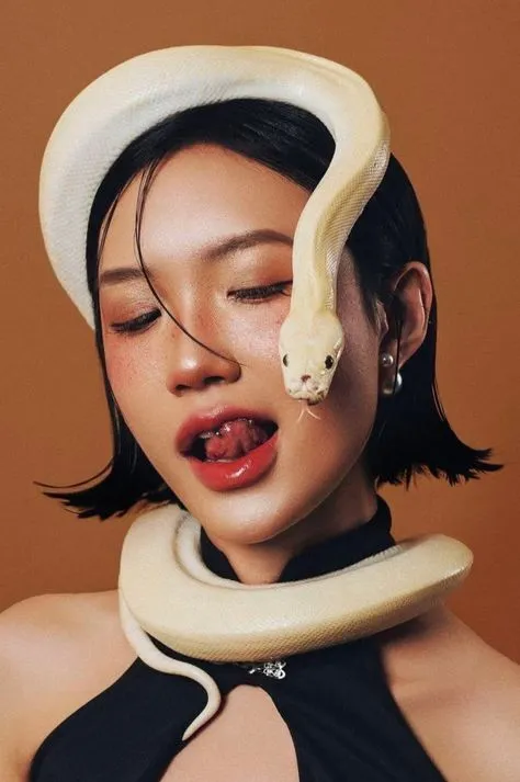 a woman with a snake on her head wearing a black dress and white hat is posing for the camera