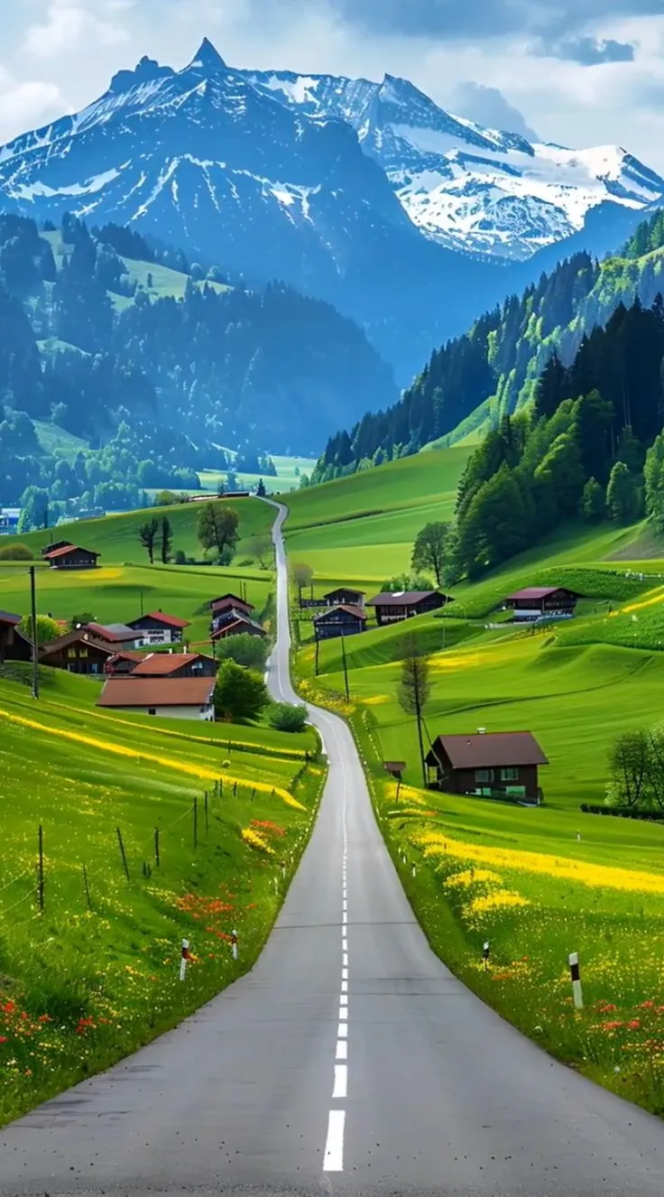 an empty road in the middle of a green valley with houses and mountains behind it